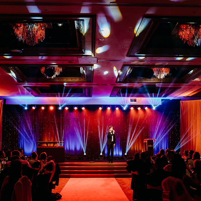 A person stands on a brightly lit stage with colorful spotlights and chandeliers overhead, addressing an audience seated at tables—a scene expertly crafted by a leading event production company in Auckland.