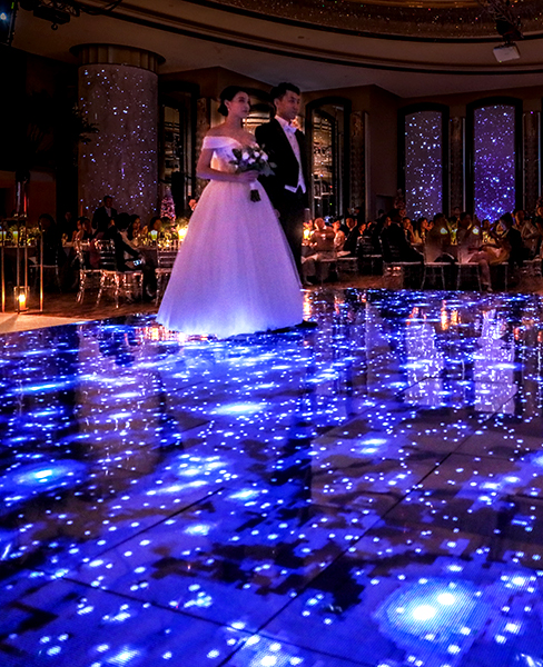 A bride and groom walk together on a dance floor illuminated with bright, star-like blue lights, surrounded by seated guests in a warmly lit, elegant weddings venue.