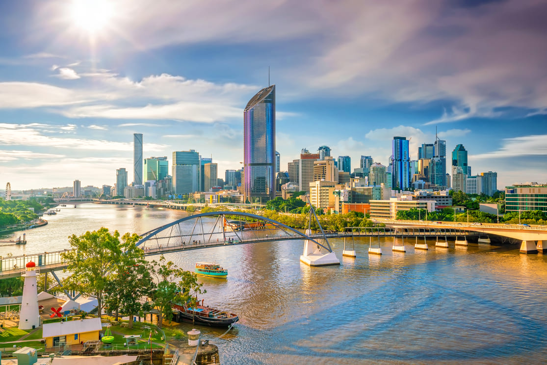 A vibrant city skyline with modern high-rise buildings along a river, a bridge spanning the water, boats docked at the riverbank, and the sun shining brightly in a partly cloudy sky.