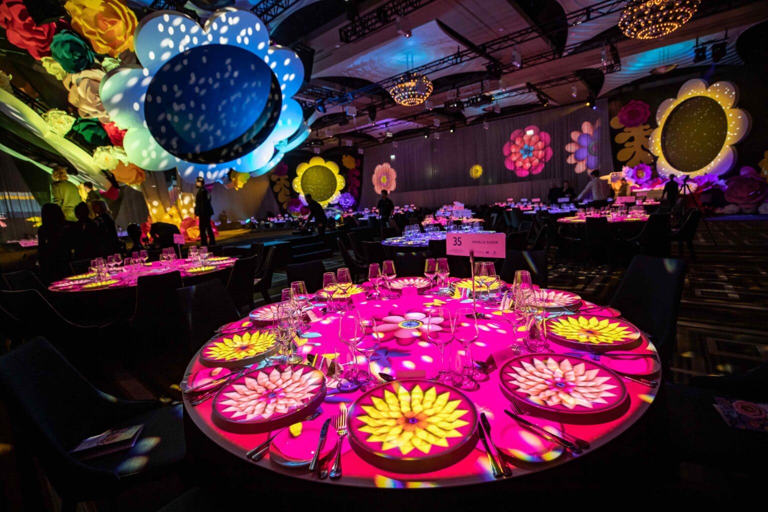 A vibrant event space with round tables illuminated by colorful, flower-shaped projections. Giant, decorative flowers and soft, colorful lighting fill the room, creating a whimsical, festive atmosphere. Table 35 is visible in the foreground.