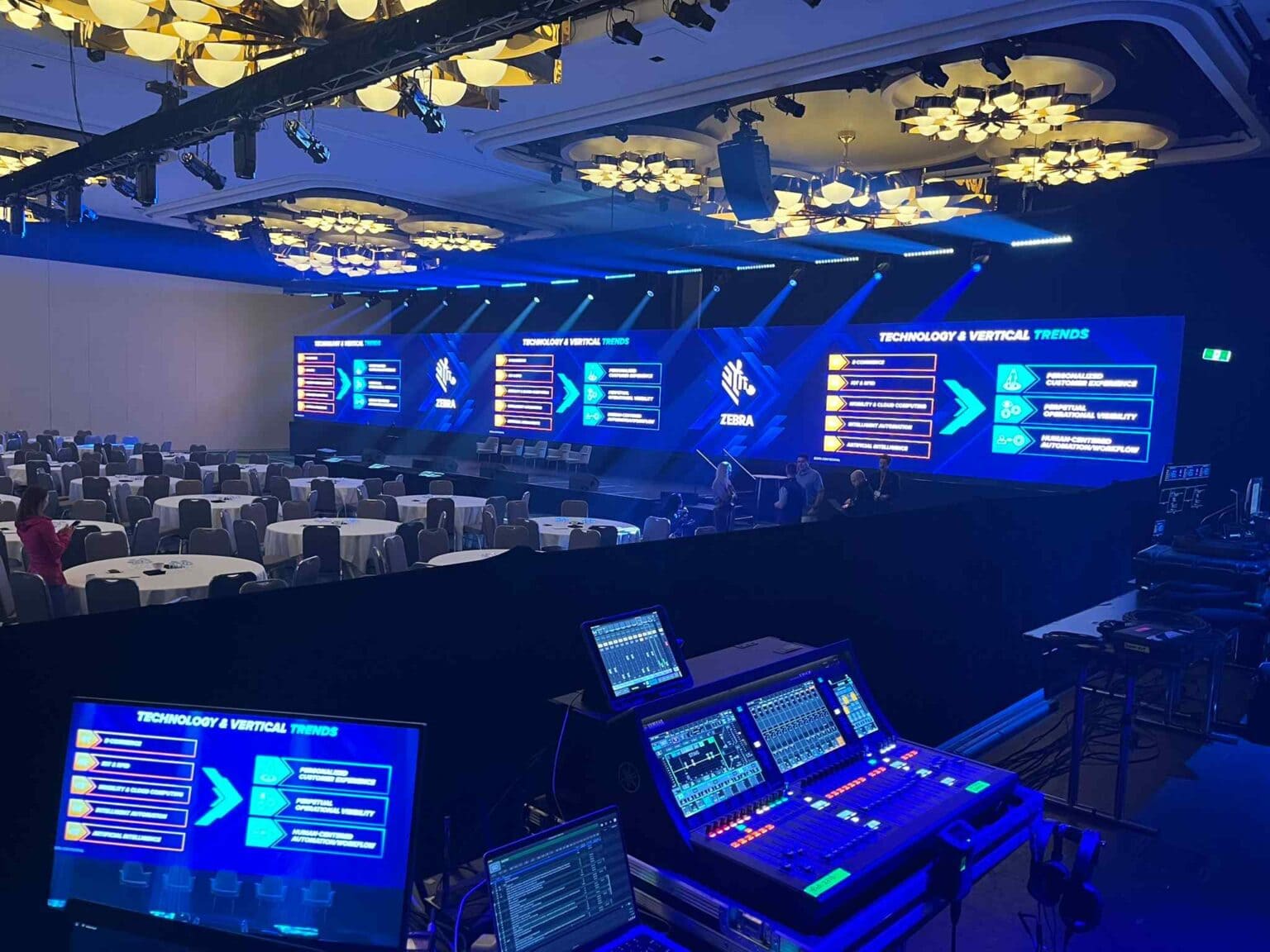 A large conference room with round tables and chairs, featuring a wide stage with multiple large LED screens displaying technology and vertical trend topics. Audio-visual control equipment is set up in the foreground.