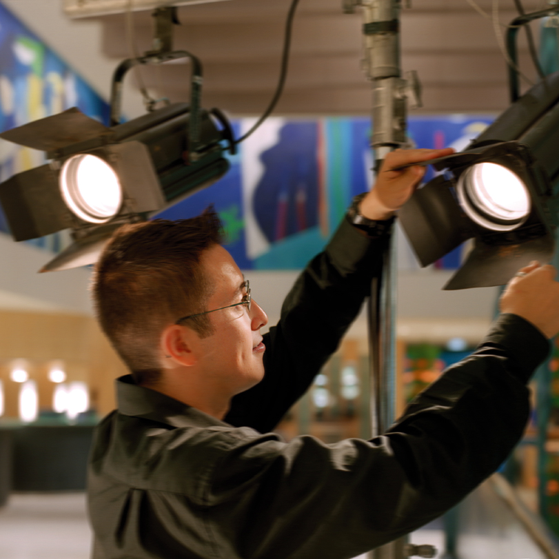 A person in glasses adjusts a stage spotlight indoors, surrounded by colorful artwork and modern decor in the background.