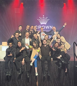 A large group of people pose together on a stage, smiling and making playful gestures in front of a Crown event backdrop under red and purple lighting—a perfect snapshot of why work with Encore is both fun and rewarding.