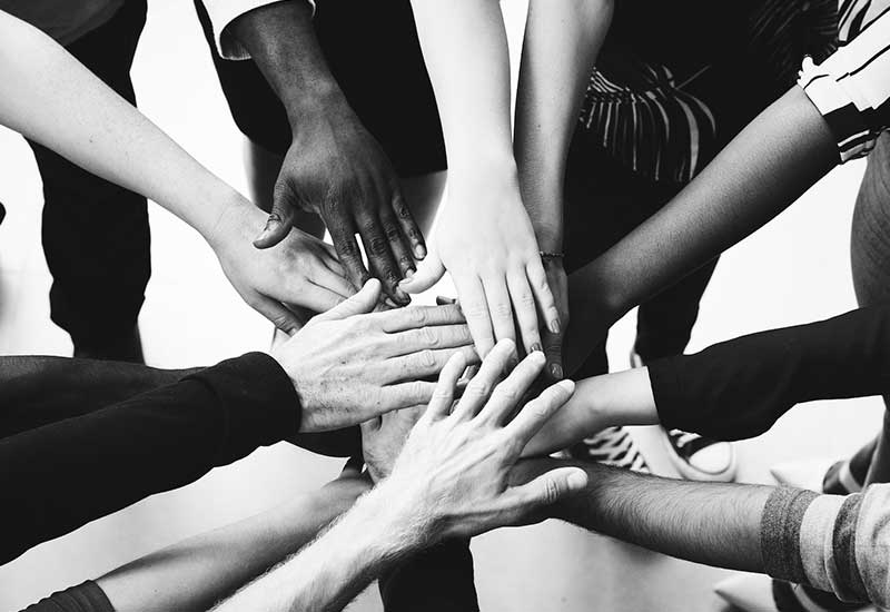 A group of diverse people standing in a circle place their hands together in the center, symbolizing unity, teamwork, and collaboration. The image is in black and white.