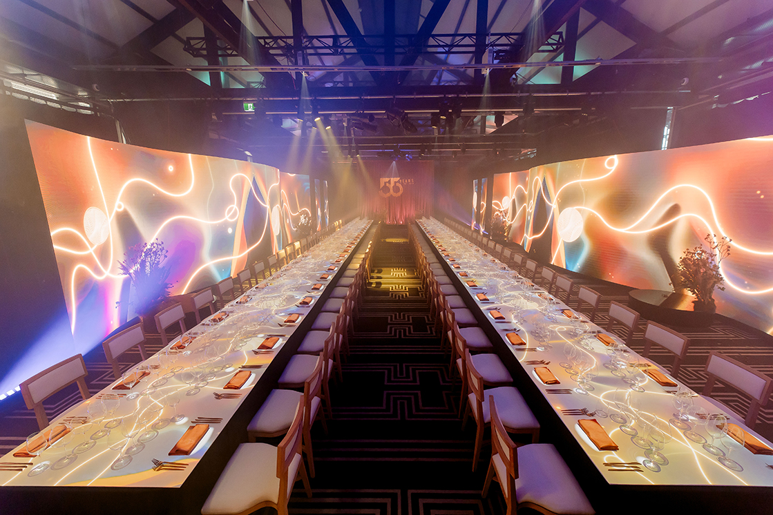 A lavish event setup with two long banquet tables, elegant place settings, and dramatic lighting. Large, colorful digital screens with abstract designs flank the tables, creating a festive, upscale atmosphere.