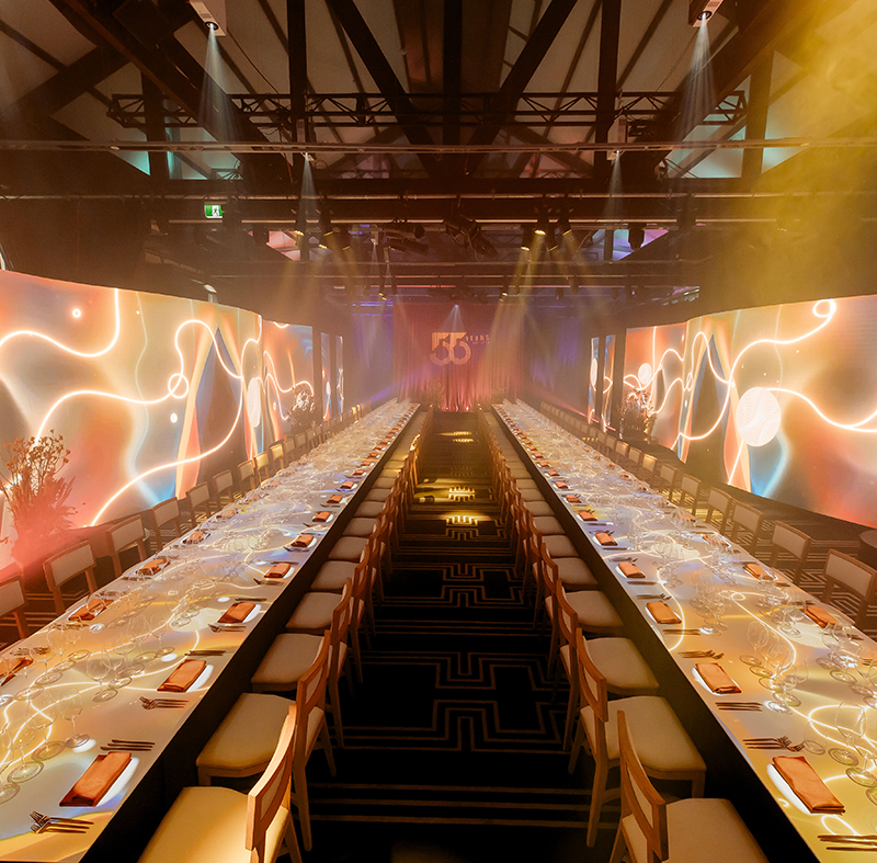 A long, elegant banquet table set for an event by Encore Sydney, with modern lighting, decorative plates and glasses, surrounded by wooden chairs. Colorful, abstract light projections by this leading AV company Sydney illuminate the walls and table surface.