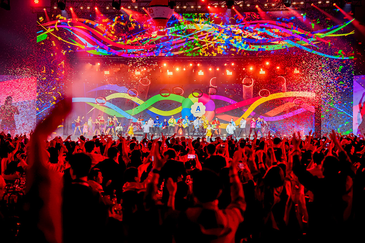 A large crowd cheers and applauds at a vibrant indoor concert with colorful lights, confetti, and performers on a stage set for gala dinner. Bright, multicolored waves decorate the backdrop, creating a festive atmosphere.