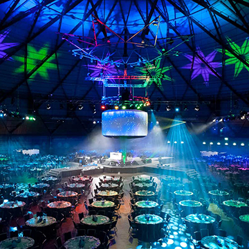 A large event venue with round tables set for guests, colorful stage lighting, and a central stage with screens displaying blue star patterns under a dome ceiling.