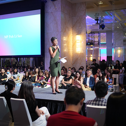 A woman in a green dress stands on stage speaking into a microphone, holding papers. Behind her, a large screen displays MP Poh Li San—an example of seamless work by an event production company in Singapore for events in Singapore.