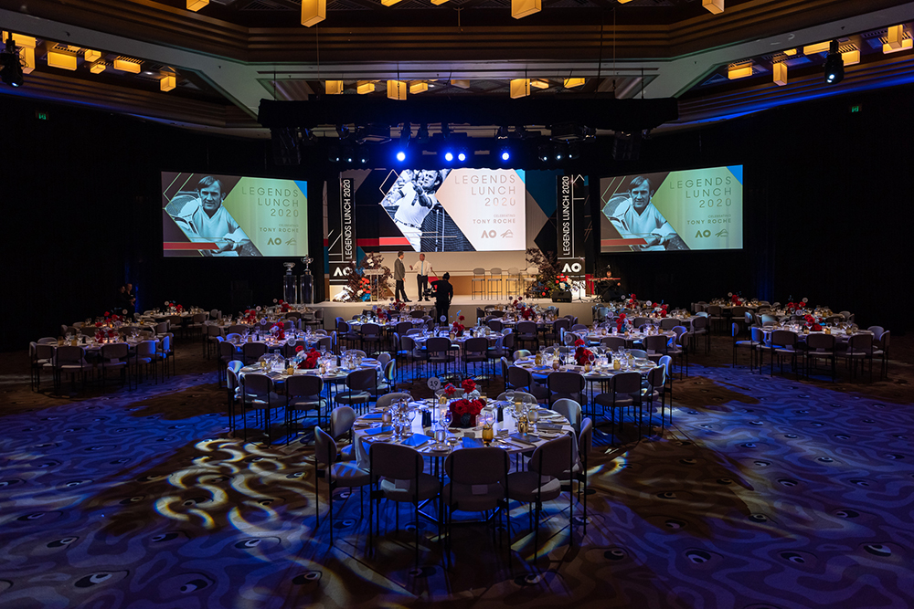 Elegant banquet hall set for an event, with round tables and floral centerpieces, stage at the front, large screens displaying Legends Lunch 2024 and images of tennis players, and dramatic blue lighting.