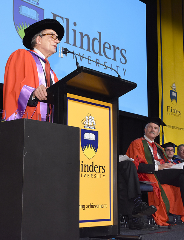 A man in academic robes and a cap speaks at a podium with the Flinders University logo, while other officials in robes sit on stage in the background. A large Flinders University sign is displayed behind him.