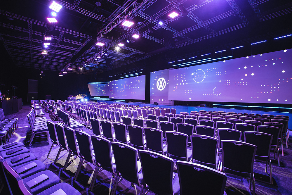 Rows of empty chairs face a large stage with wide digital screens displaying abstract graphics and the Volkswagen logo, illuminated by purple and blue lighting in a spacious venue, perfect for hosting a conference and gala dinner.