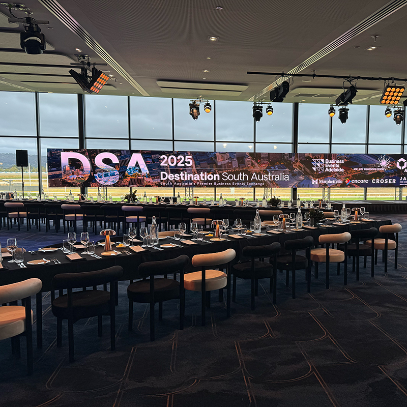 A modern conference room with large windows, set up for an event by Encore Adelaide, features long tables with place settings and a large digital screen displaying DSA 2025 Destination South Australia.