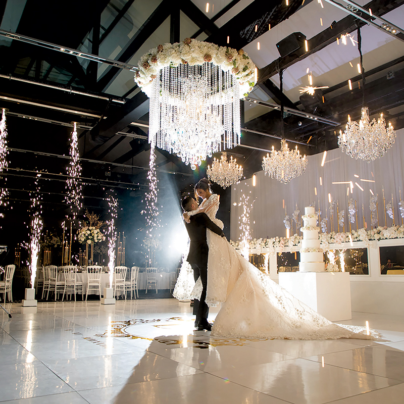 A bride and groom share their first dance under a sparkling chandelier at their dream wedding, surrounded by fireworks, elegant decor with white flowers, and a large tiered cake in the background.