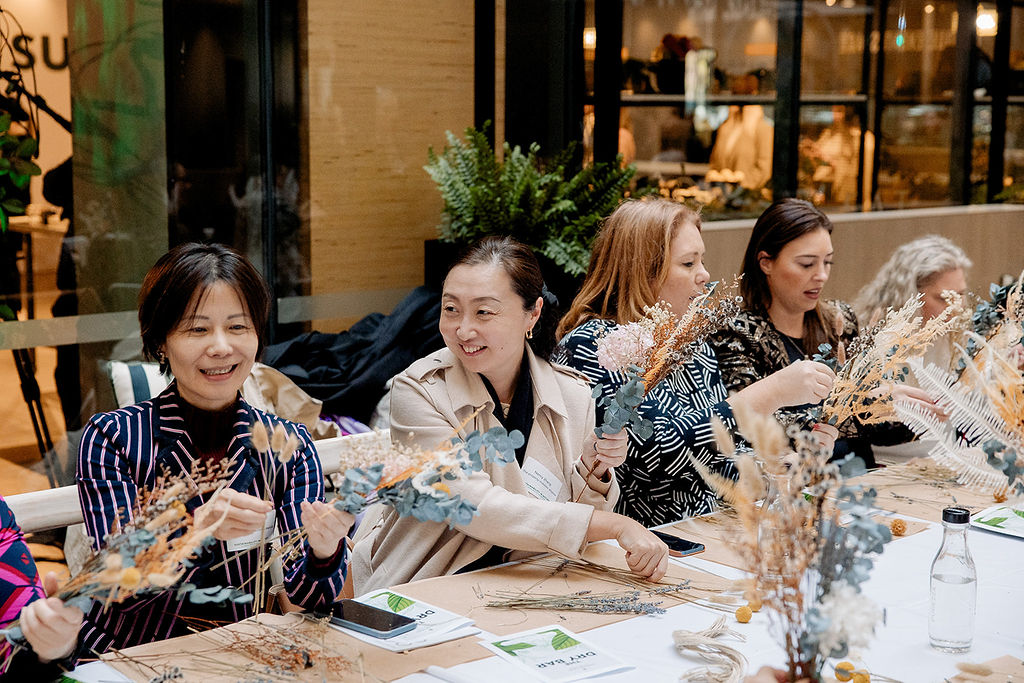 Five women sit at a table, smiling and arranging dried flowers during a workshop. The table is covered with floral materials and paperwork, and a green plant is visible in the background.