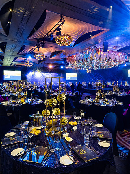 A glamorous, dimly lit banquet hall decorated with gold centerpieces and sparkling chandeliers, set for a formal event with round tables, elegant place settings, and a stage with bright lights in the background.