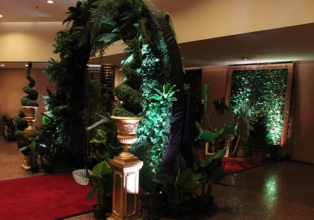 A decorative indoor archway covered in lush green foliage and plants, lit with green spotlights, stands on a red carpet, flanked by gold planters and additional potted plants.