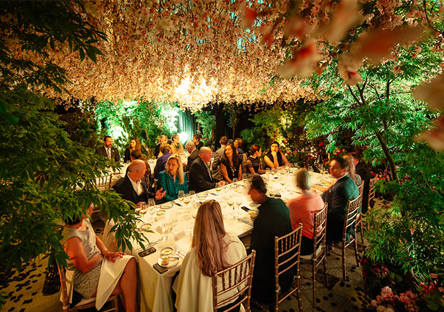 Guests sit around two long, elegantly set dining tables surrounded by lush greenery and trees, with pink and white flowers hanging overhead, creating a magical, garden-like atmosphere at an indoor event.