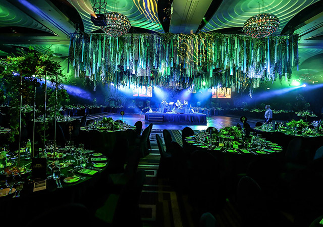 A lavish, green-lit banquet hall with round tables set for dining, featuring an elevated stage where musicians perform. Streamers and foliage hang from the ceiling, and chandeliers add a touch of elegance.