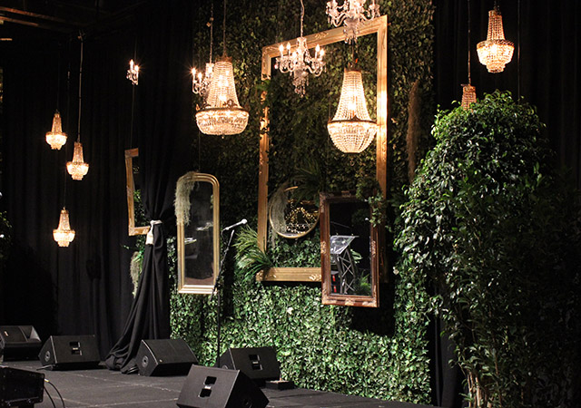 A stage decorated with lush green foliage, gold-framed mirrors, and chandeliers hanging above. Microphones and speakers are set up, with black curtains framing the space, creating an elegant and lush atmosphere.