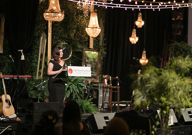 A woman in a black dress speaks at a podium with a Fight Cancer Foundation sign, surrounded by greenery, chandeliers, and string lights at an elegant indoor event.