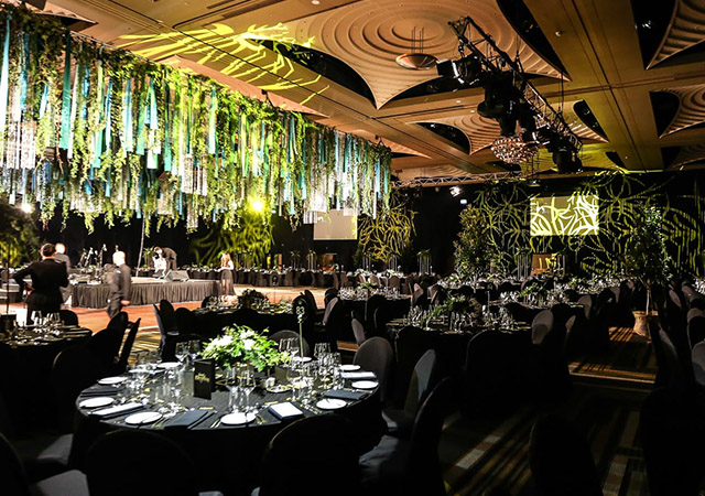 A large, elegant banquet hall decorated with hanging greenery and plants from the ceiling. Round tables are set with glassware and plates, and stage lighting creates a warm, festive atmosphere.
