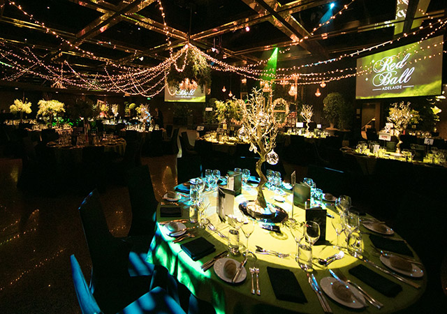 Elegant ballroom set for an event, with round tables covered in green cloths, formal place settings, and centerpiece decorations. Overhead, string lights and chandeliers create a warm, festive atmosphere. Screens display “Red Ball Adelaide.”.