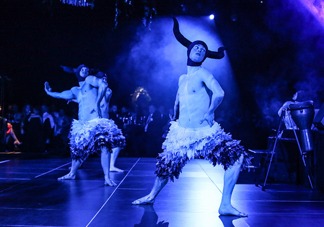 Two performers wearing horned headpieces and feathered skirts dance dramatically on a dimly lit stage with blue lighting and smoke, as an audience watches in the background.