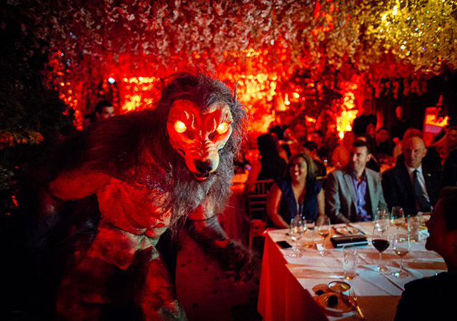A person in a detailed werewolf costume with glowing red eyes walks past a group of people seated at a dinner table in a dimly lit, elaborately decorated room with red lighting and hanging flowers.