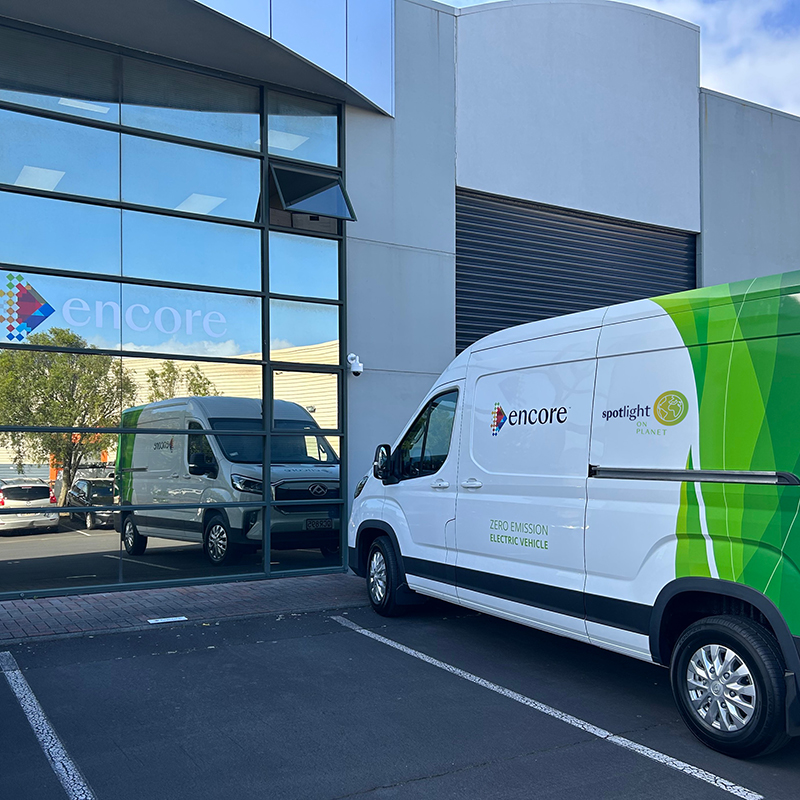 Two white vans are parked outside a modern office building with large windows. The van in front is branded with encore and Spotlight Planet logos and labeled as a zero emission electric vehicle. The buildings sign reads encore.