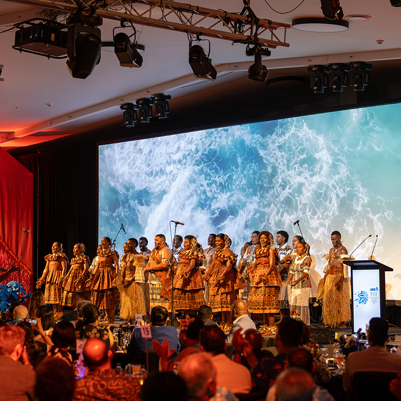A group of performers in traditional attire sing on stage with an ocean backdrop, entertaining guests at round tables during an indoor event—expertly managed by a leading event production company in Fiji.