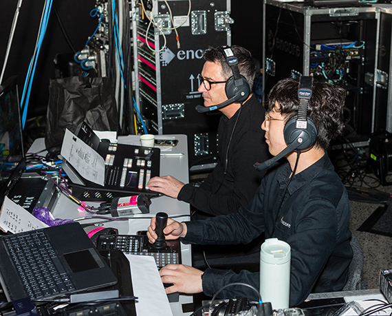Two people wearing headsets sit at a control desk with monitors, papers, and control panels, smoothly operating technical equipment backstage—showing why work with Encore means partnering with dedicated event professionals.