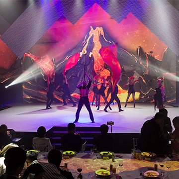 Performers dance on stage with dramatic red fabric and a fiery volcano backdrop, while an eager audience enjoys food and drinks. Bright stage lights, swirling smoke, and calls for an encore create a dynamic atmosphere.