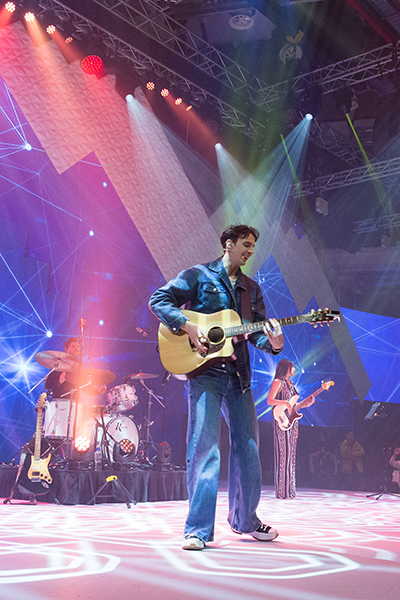 A musician in a denim jacket and jeans plays an acoustic guitar on a brightly lit stage, with a drummer and bassist performing in the background amid colorful lights and geometric projections.