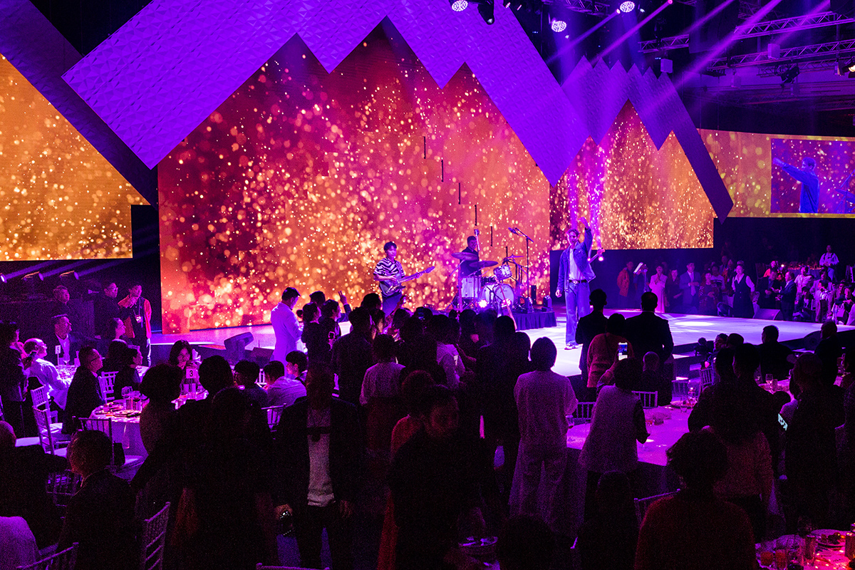 A live band performs on a brightly lit stage set for a gala dinner, featuring gold and purple light displays, while a large audience sits and stands around tables at this formal event.