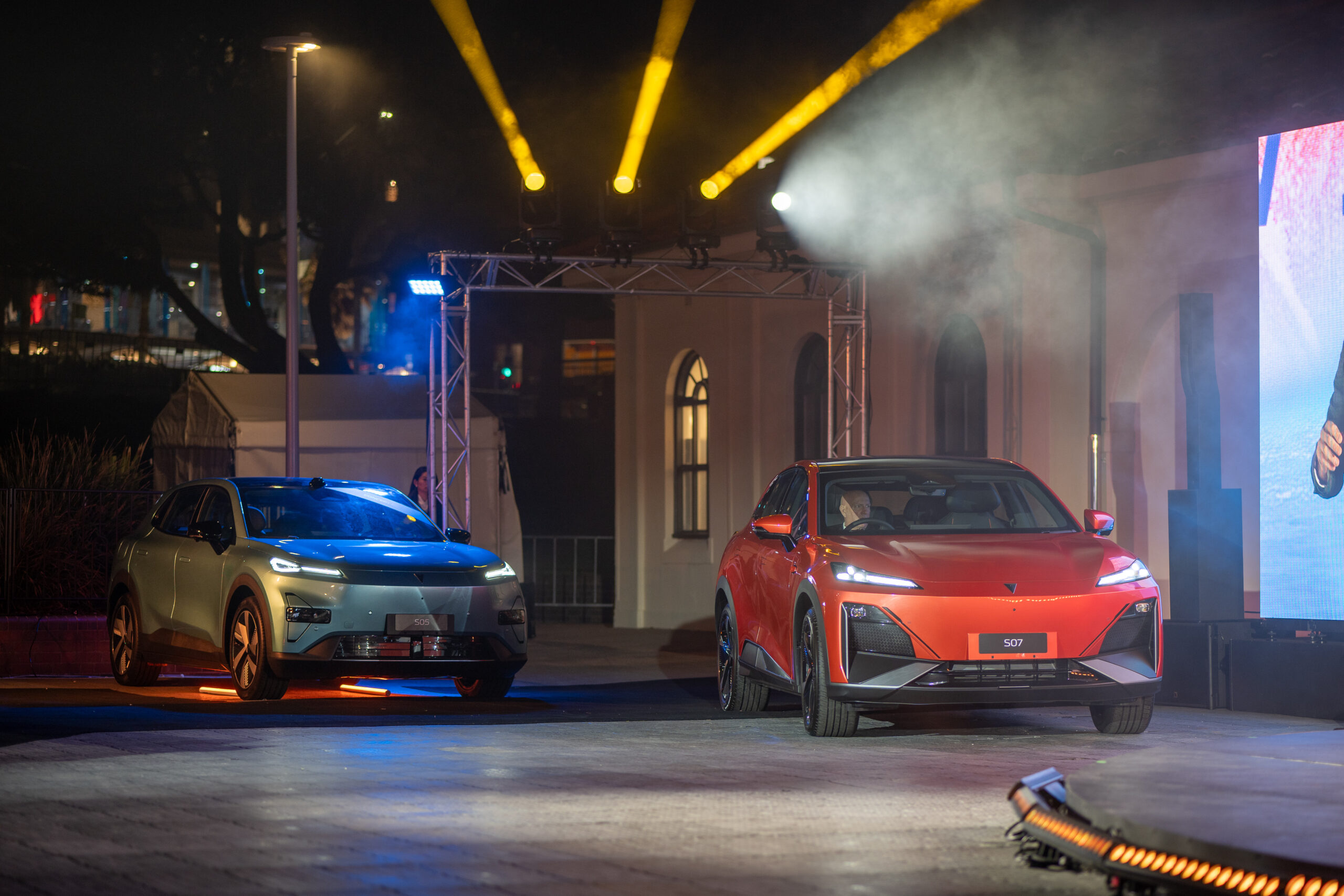 Two modern electric SUVs, one silver and one red, are showcased on an outdoor stage with theatrical lighting and fog effects, highlighting their sleek designs at a nighttime event.