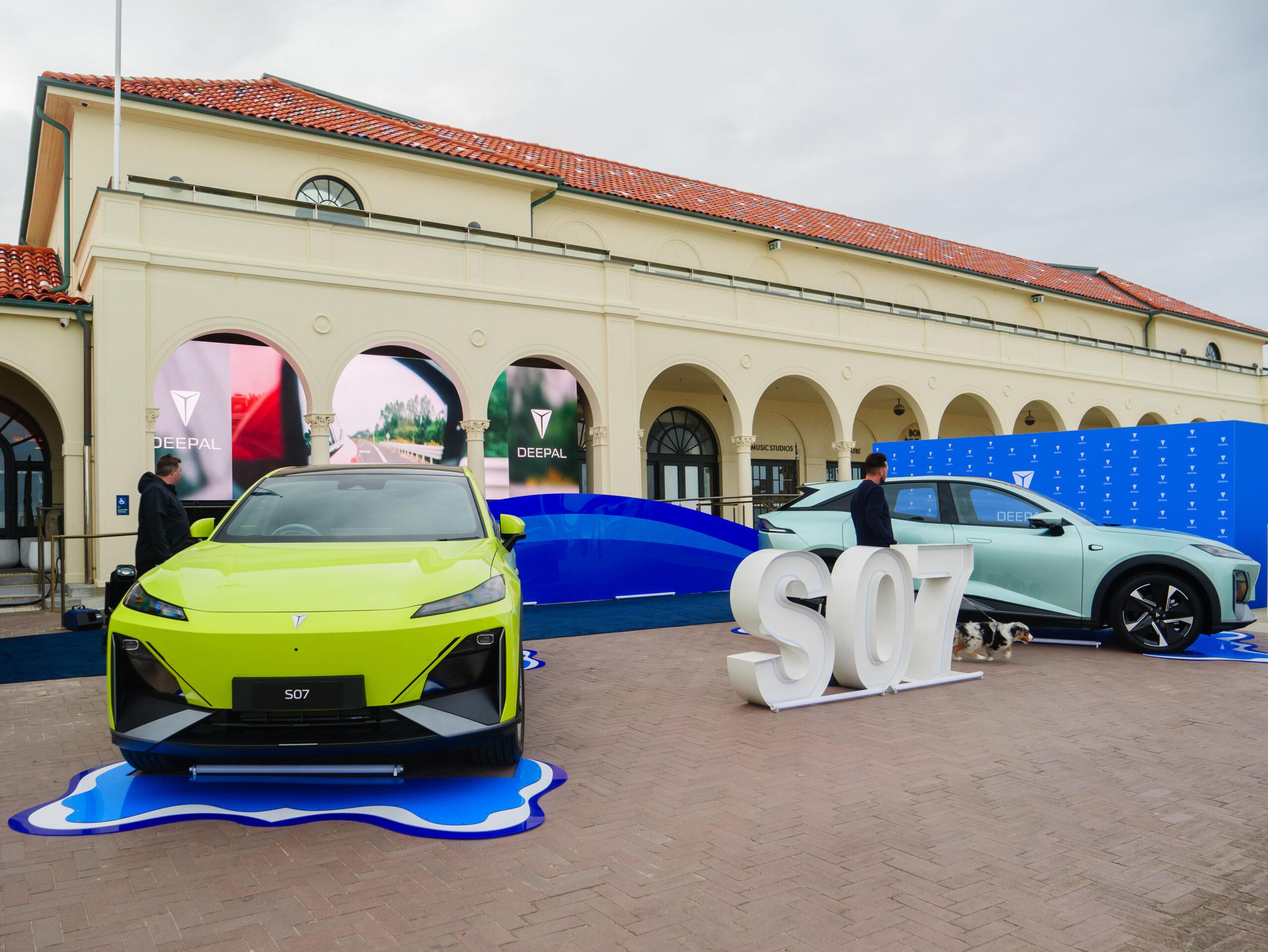 Two modern electric cars, one bright green and one light blue, are displayed outdoors in front of an elegant building with arches and red-tiled roof. Large S07 letters and DEEPAL branding are visible.