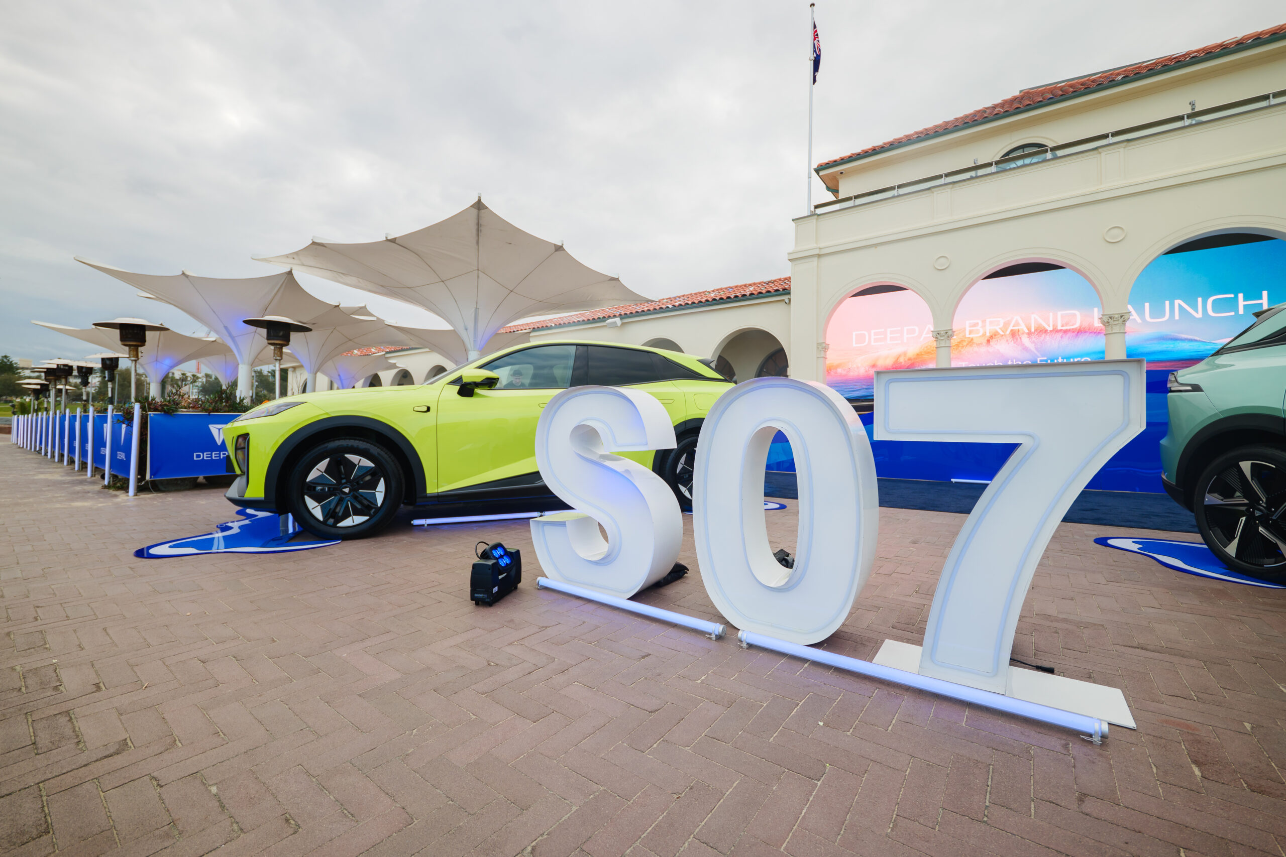 A bright yellow car is parked near large white letters spelling S07 outside an event venue with a DEEPAL BRAND LAUNCH sign and white canopy structures in the background.