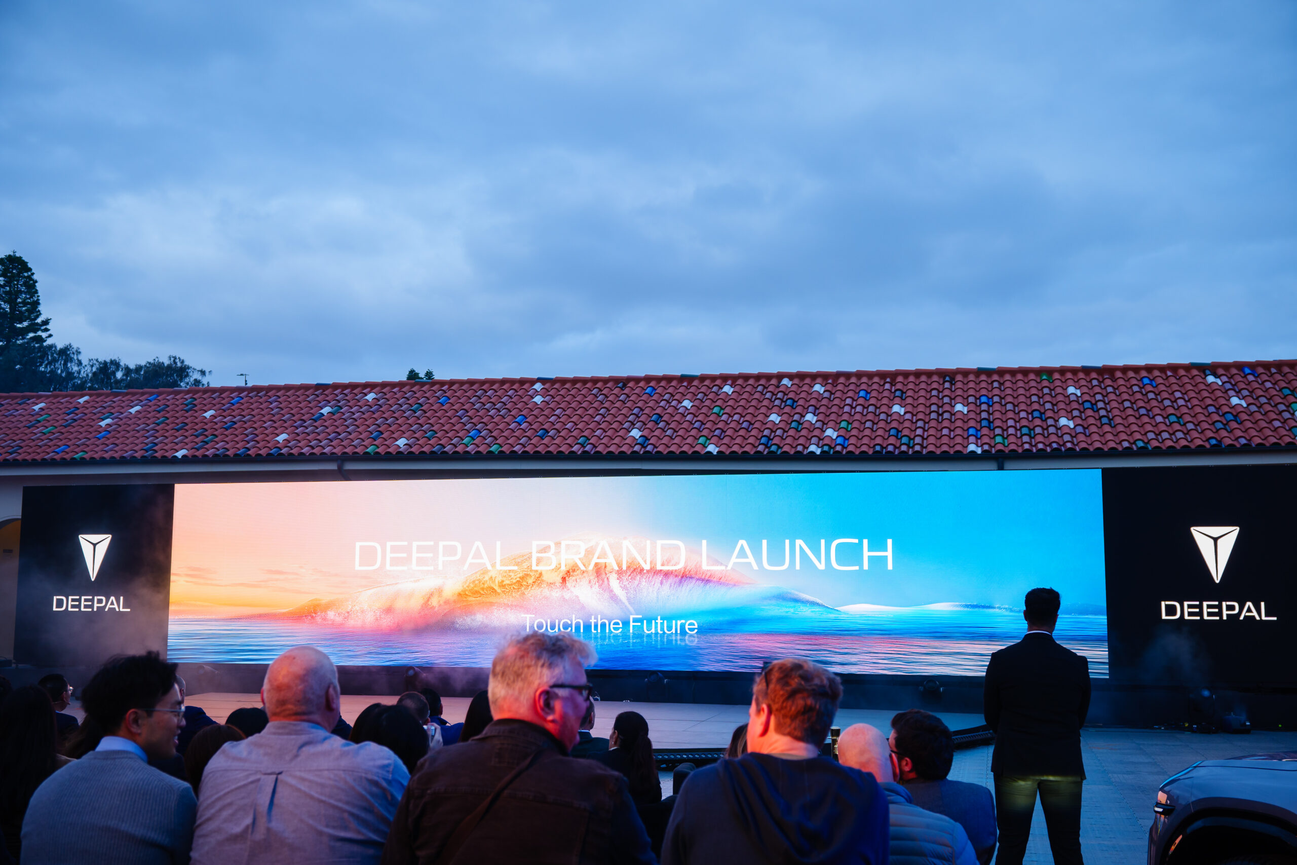 An audience sits facing a large, colorful digital screen displaying DEEPAL BRAND LAUNCH and Touch the Future at an outdoor event, with cloudy skies and a tiled-roof building in the background.
