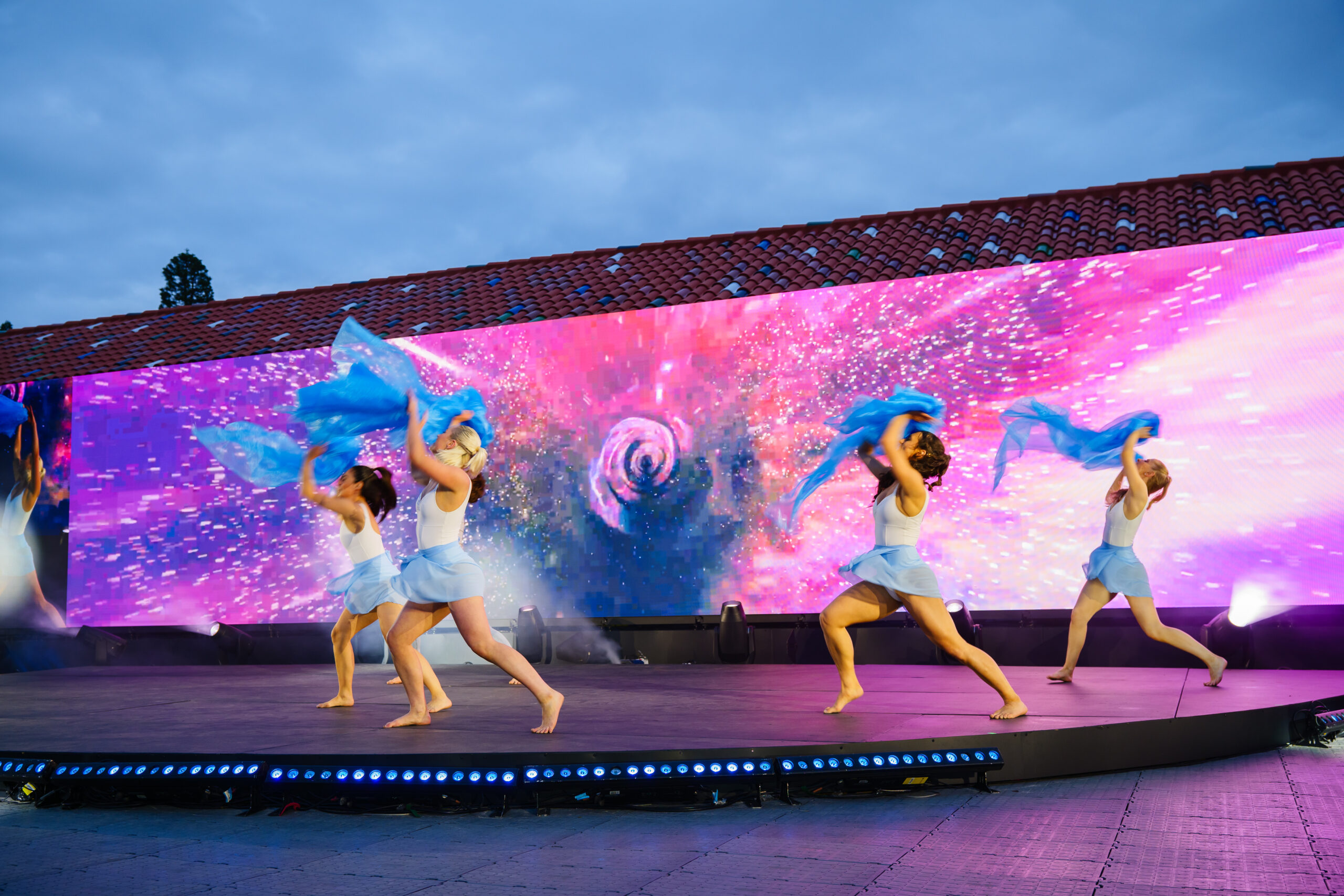Four dancers in light blue costumes perform energetically on an outdoor stage, waving blue fabric, with a colorful, cosmic-themed digital backdrop behind them under a cloudy sky.