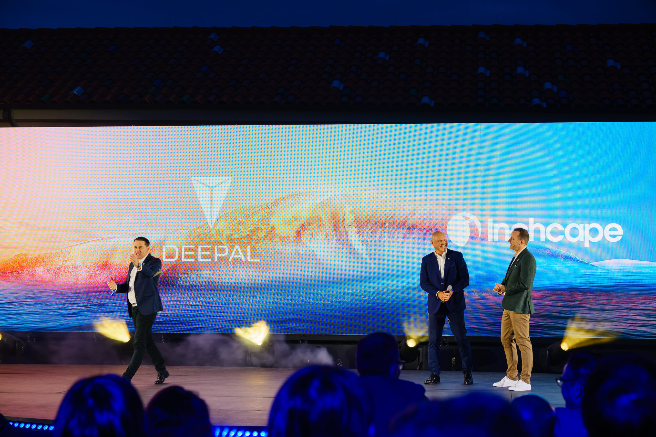 Three men stand and speak on an outdoor stage in front of a large screen displaying the Deepal and Inchcape logos, with a colorful wave graphic in the background. Audience members are visible in the foreground.