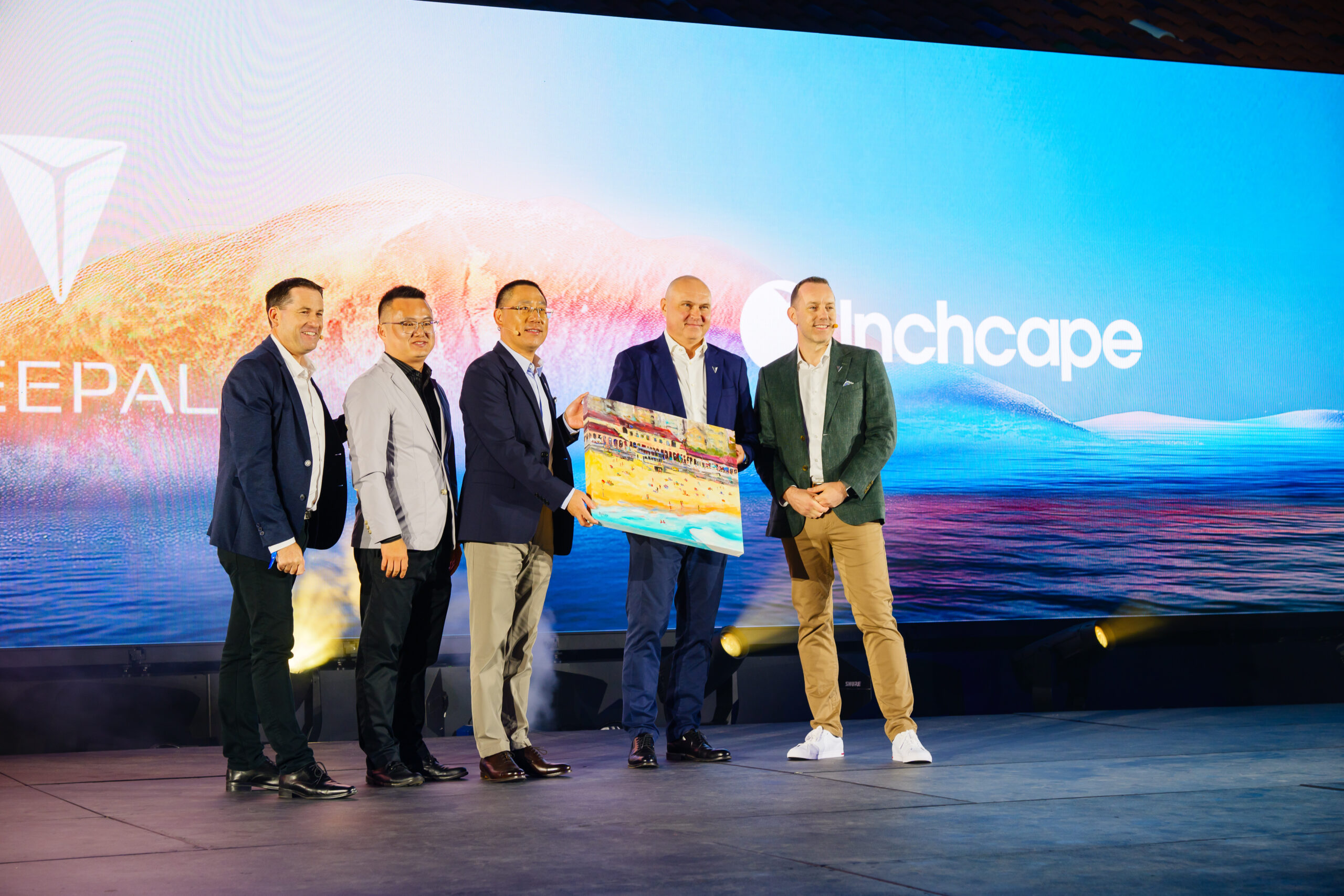 Five men in business attire stand on stage, smiling, while one hands a colorful plaque or award to another. A large screen behind them displays the logos NEPAL and Inchcape with a scenic background.