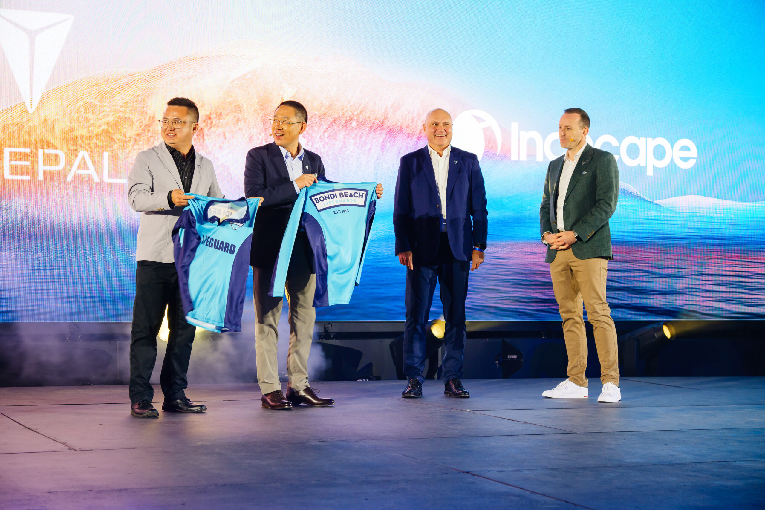 Four men stand on a stage in front of a colorful backdrop; two of them are smiling and holding up light blue sports jerseys, while the other two men stand beside them, also smiling.