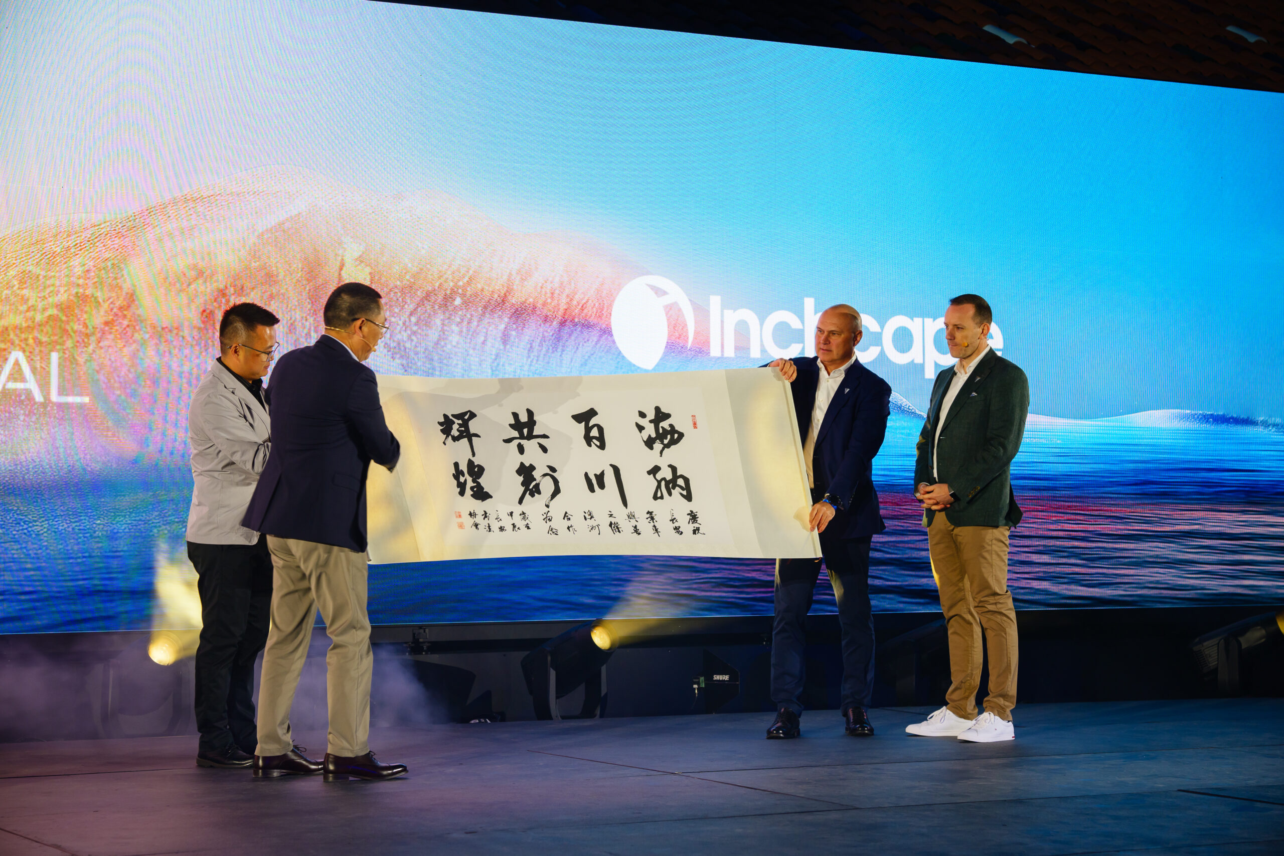 Four men in business attire stand on a stage holding a large scroll with Chinese calligraphy. A screen behind them displays the Inchcape logo and a scenic background. Stage lights and fog enhance the setting.