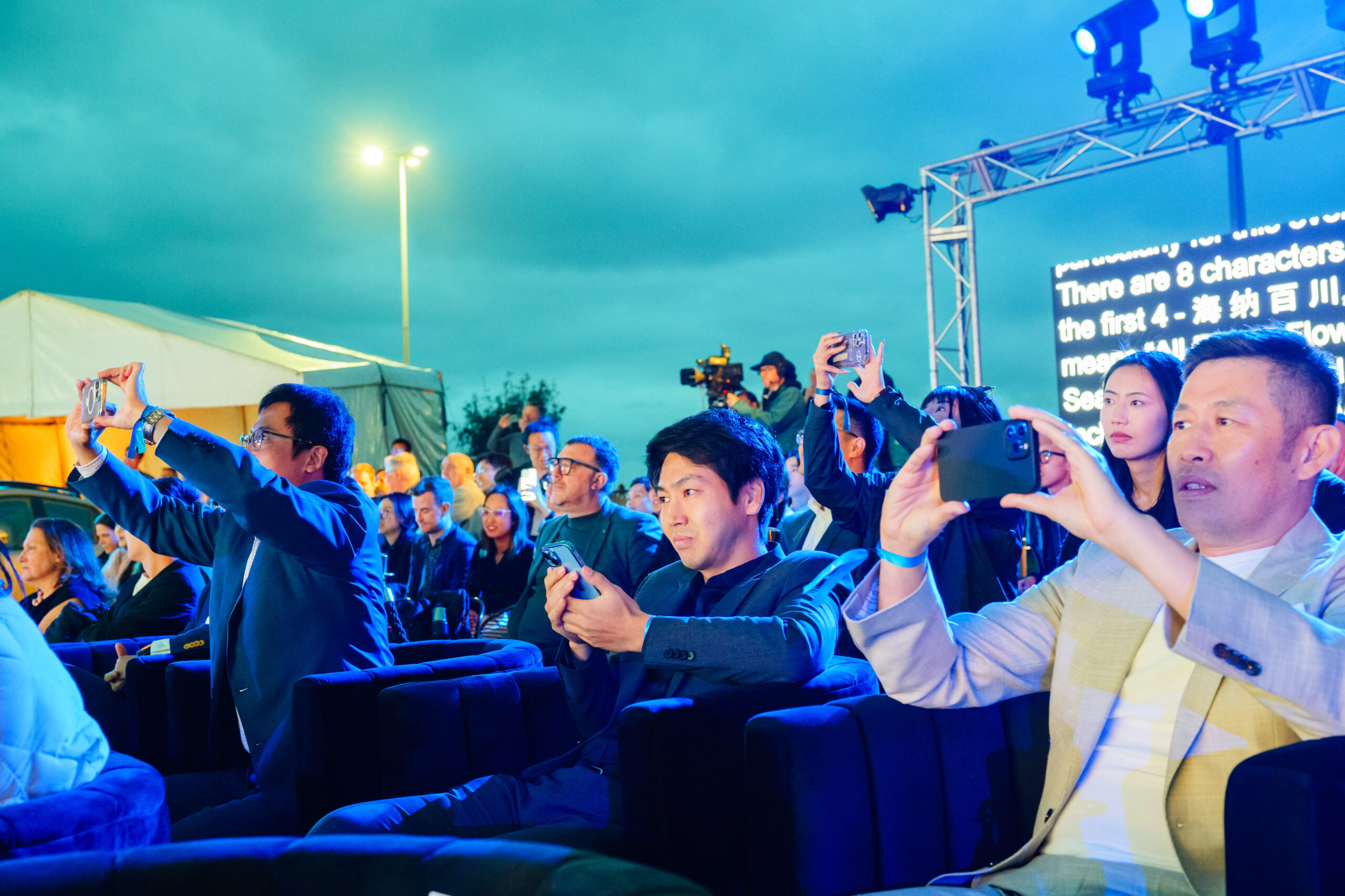 A group of people sitting in rows at an outdoor event, many holding up smartphones to take photos or videos. A teleprompter with text is visible in the background under cloudy evening skies.