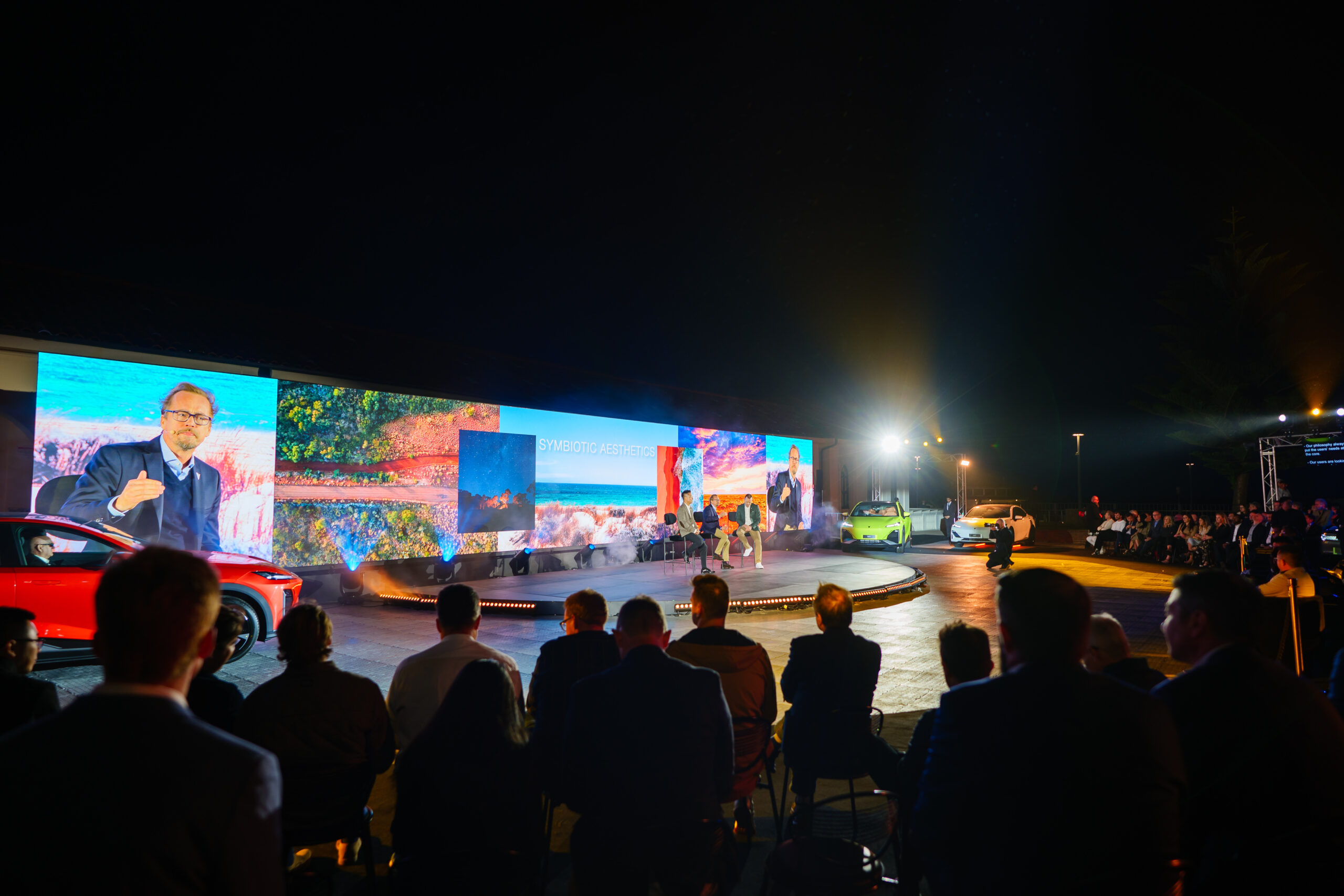 A well-lit stage at night shows a speaker addressing an audience, with large digital screens behind him displaying vibrant images and text. Several cars are on display, and attendees are seated, watching the presentation.
