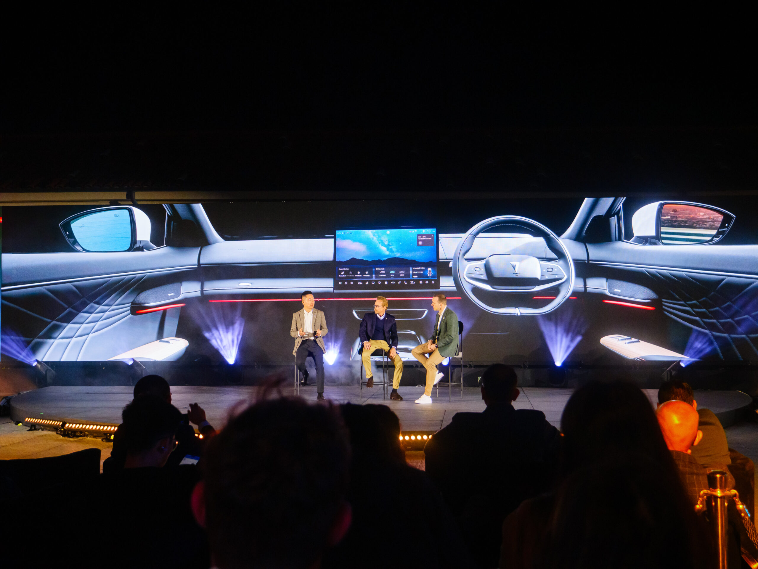 Three people sit on a stage panel discussion with a large screen behind them displaying a digital image of a cars interior dashboard and steering wheel. Audience members are seated in the foreground.