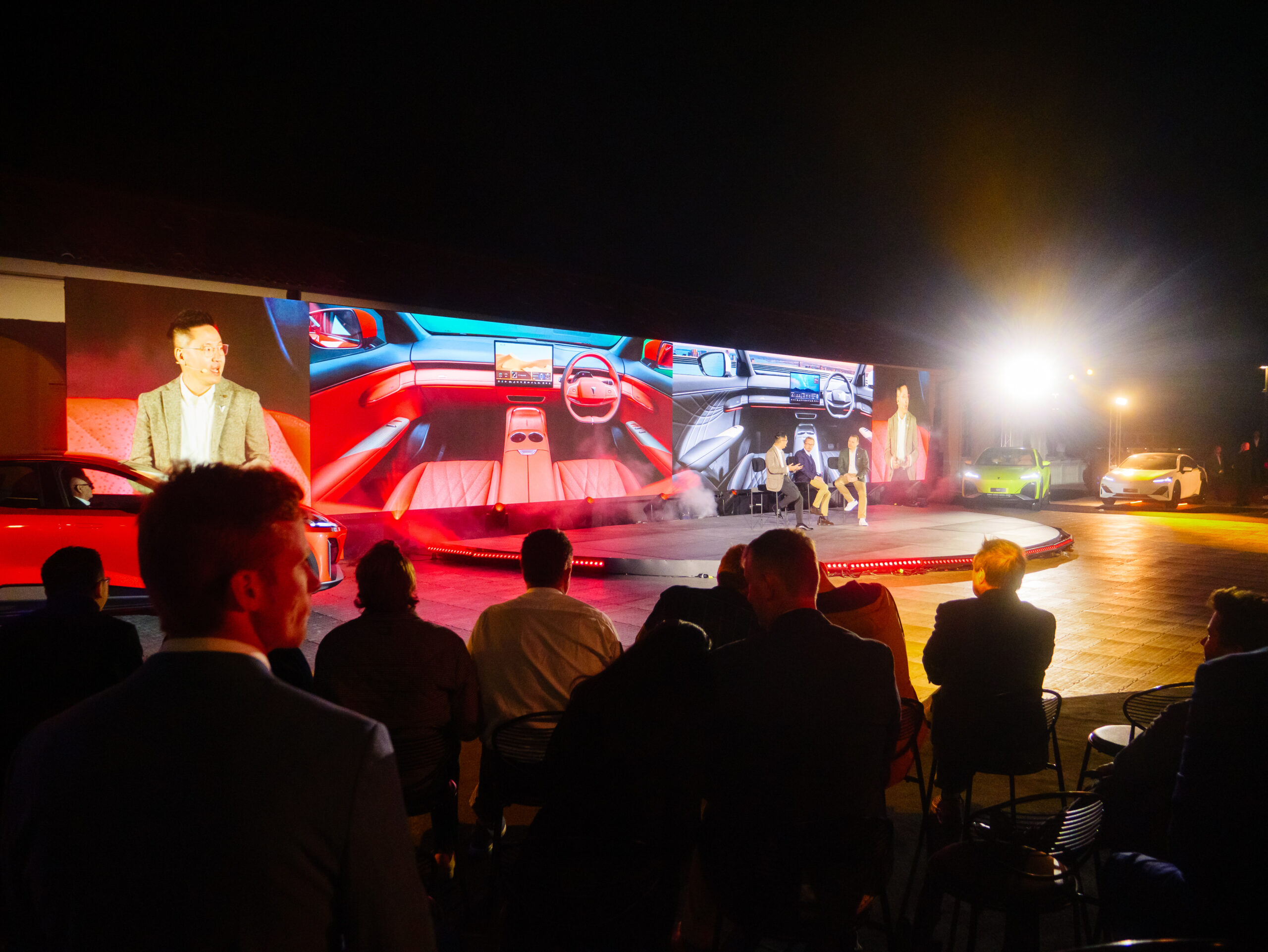 An audience watches a brightly lit outdoor presentation at night, with large screens displaying a car interior and speakers on stage. Several cars are parked nearby, and people are seated in the foreground.
