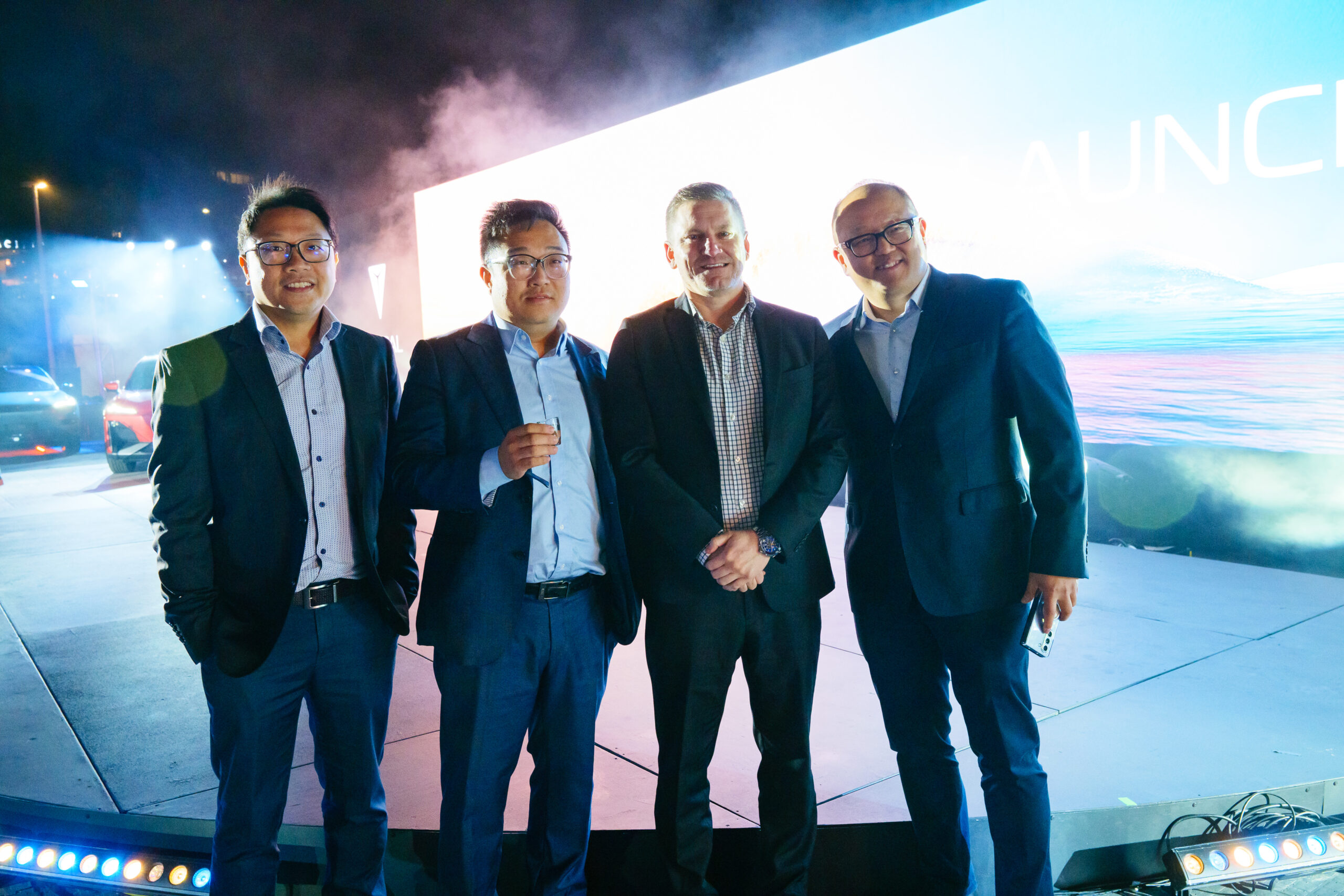 Four men in suits stand together and smile in front of a brightly lit screen at a nighttime event. Blue lights and a modern stage setup are visible in the background.