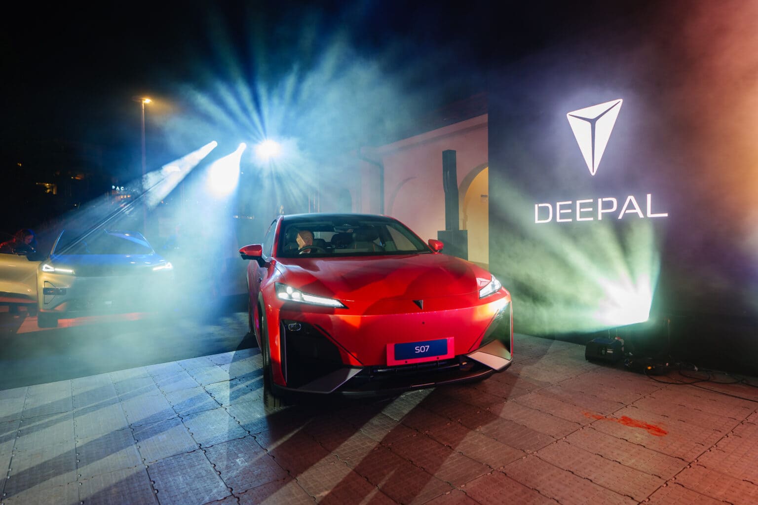 A red Deepal S07 car is showcased at night under bright spotlights, with mist and a glowing Deepal logo projected on a wall beside it. Another car is partially visible in the background.